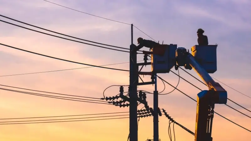 Elektrik direğinde bakım yapan işçilerin gün batımı fonunda görüntülendiği bir enerji altyapısı görseli